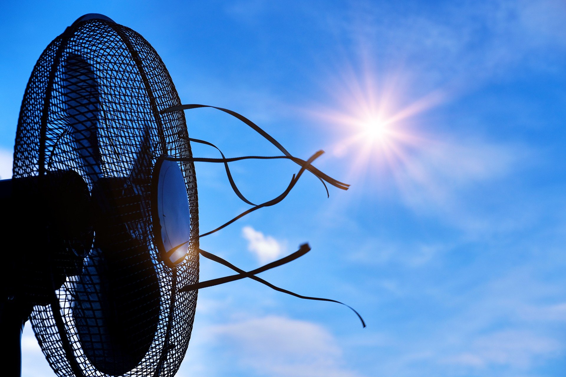 Photograph of a dark coloured electric fan facing sideways with a blue sky and sun in the background