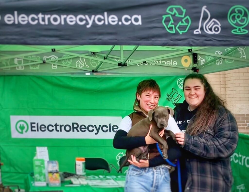Picture of Northern BC Recycling Ambassadors, Madison and Anna, at ElectroRecycle's collection booth. Madison is holding a cute puppy.
