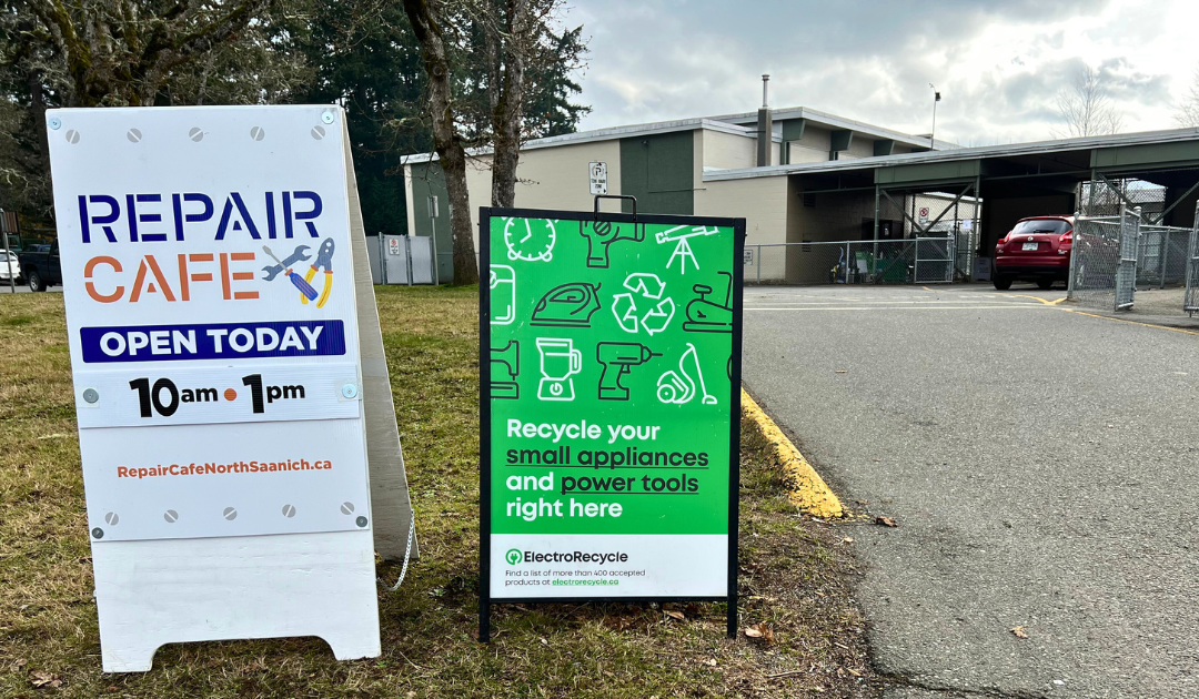 Two A-frame signs stand next to each other. On the left one that advertises 'Repair Cafe Open Today'. On the right a green sign that advertises ' Recycle your small appliances and power tools right here'.